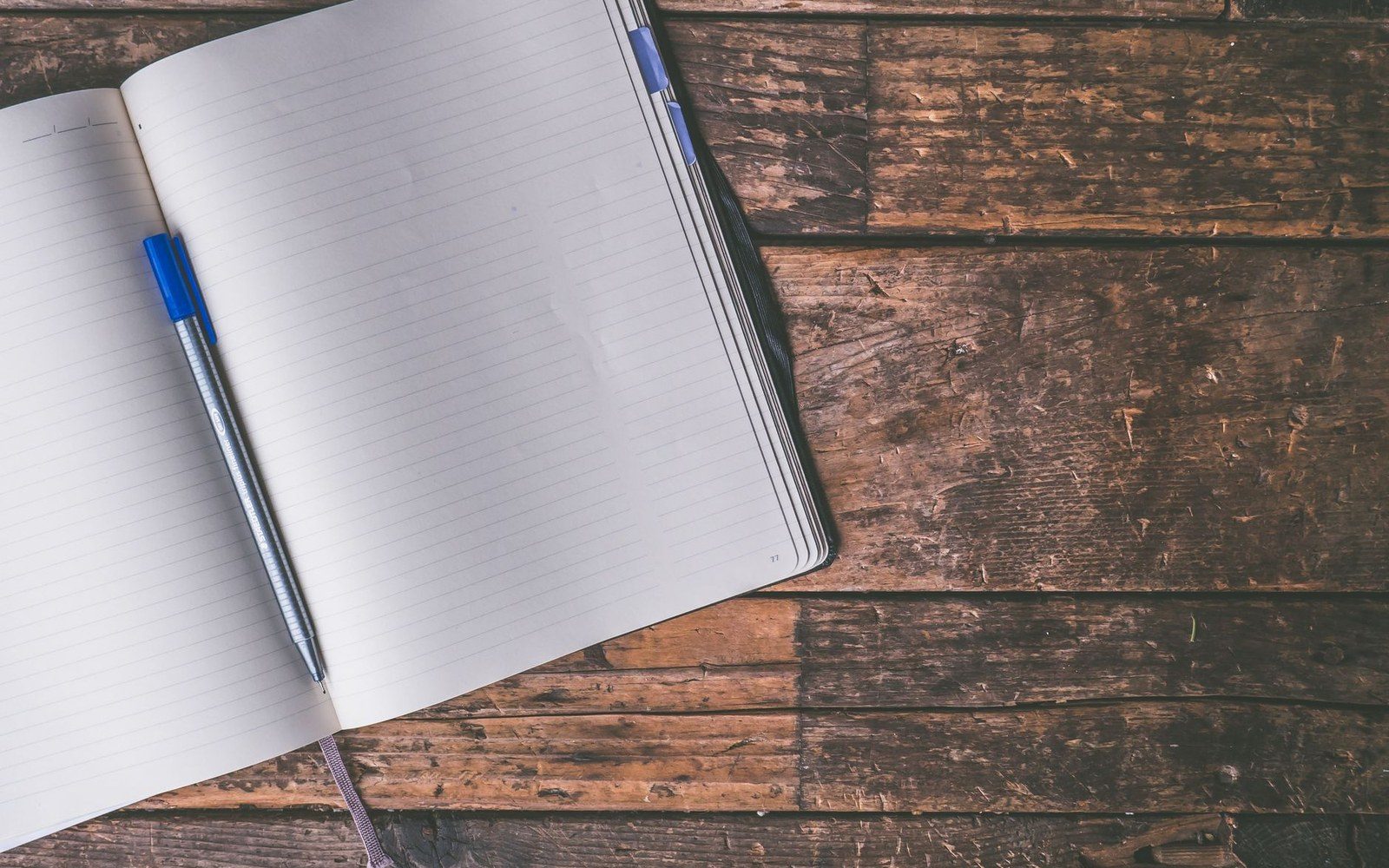 Blank lined notebook with a blue pen resting on its pages atop a wooden table