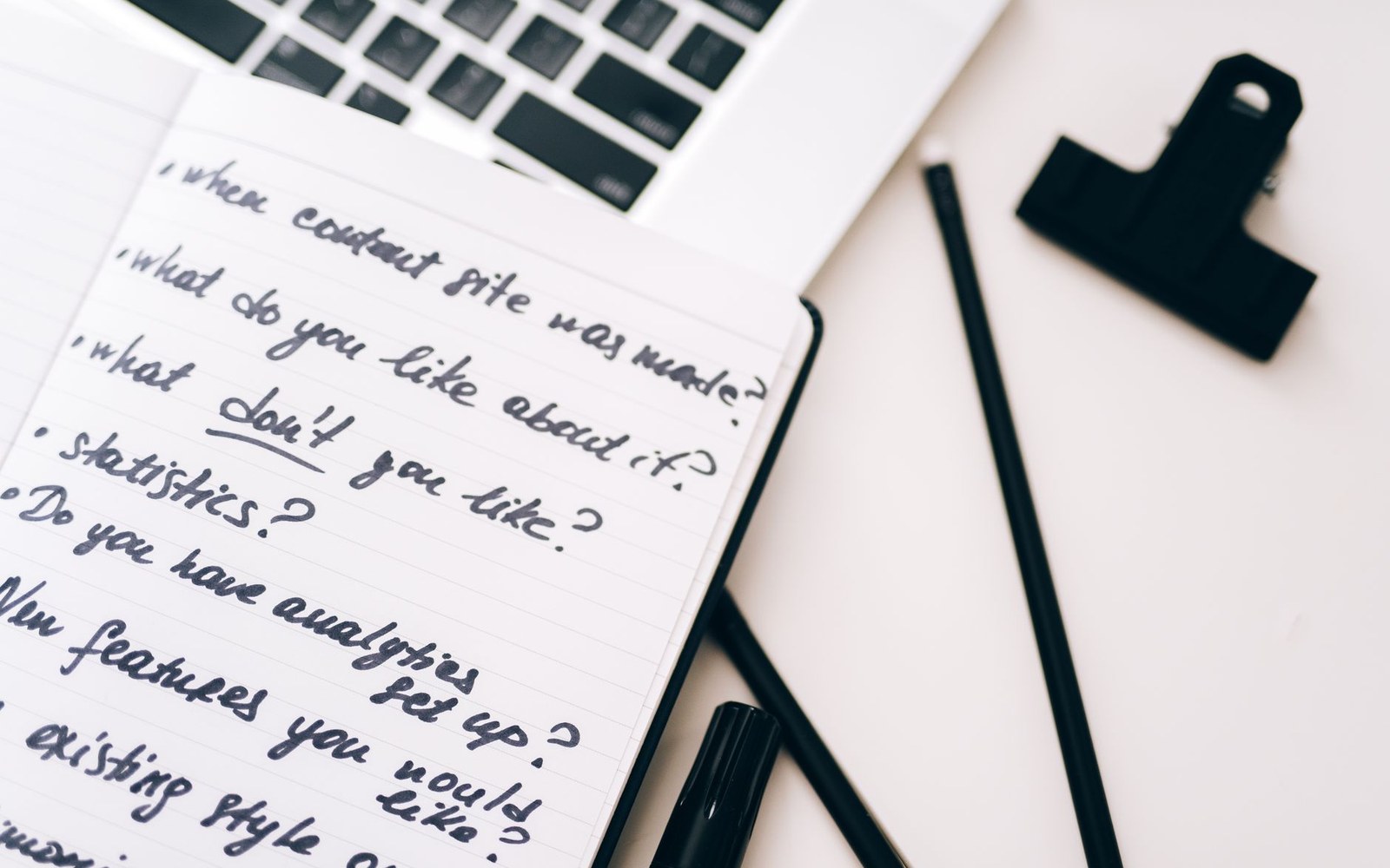 Handwritten list of interview questions in a notebook beside pens and a laptop