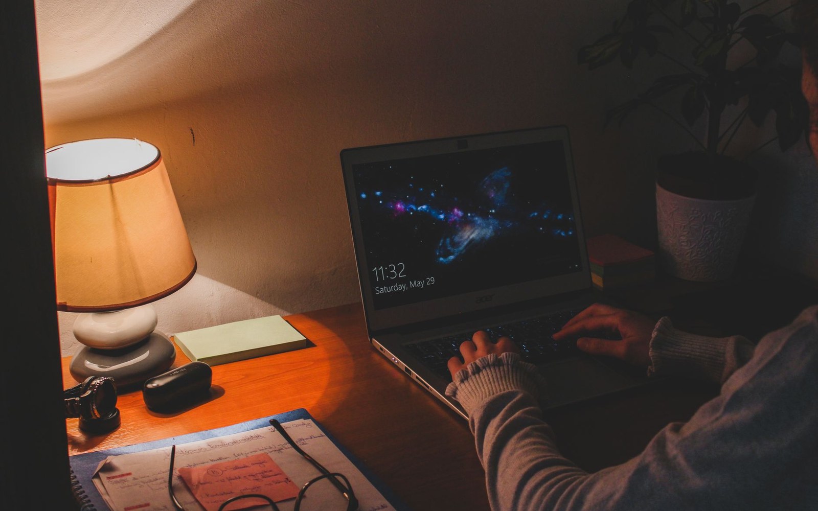 Person typing on a laptop with a nebula screensaver beside a lit desk lamp