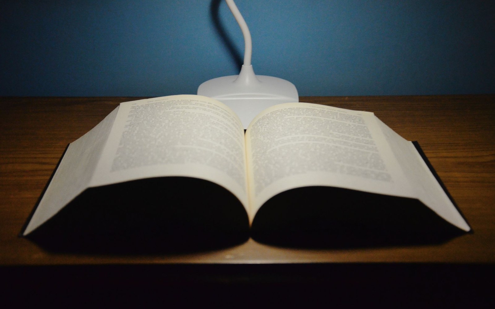 An open thick book on a wooden desk beneath a small gooseneck lamp