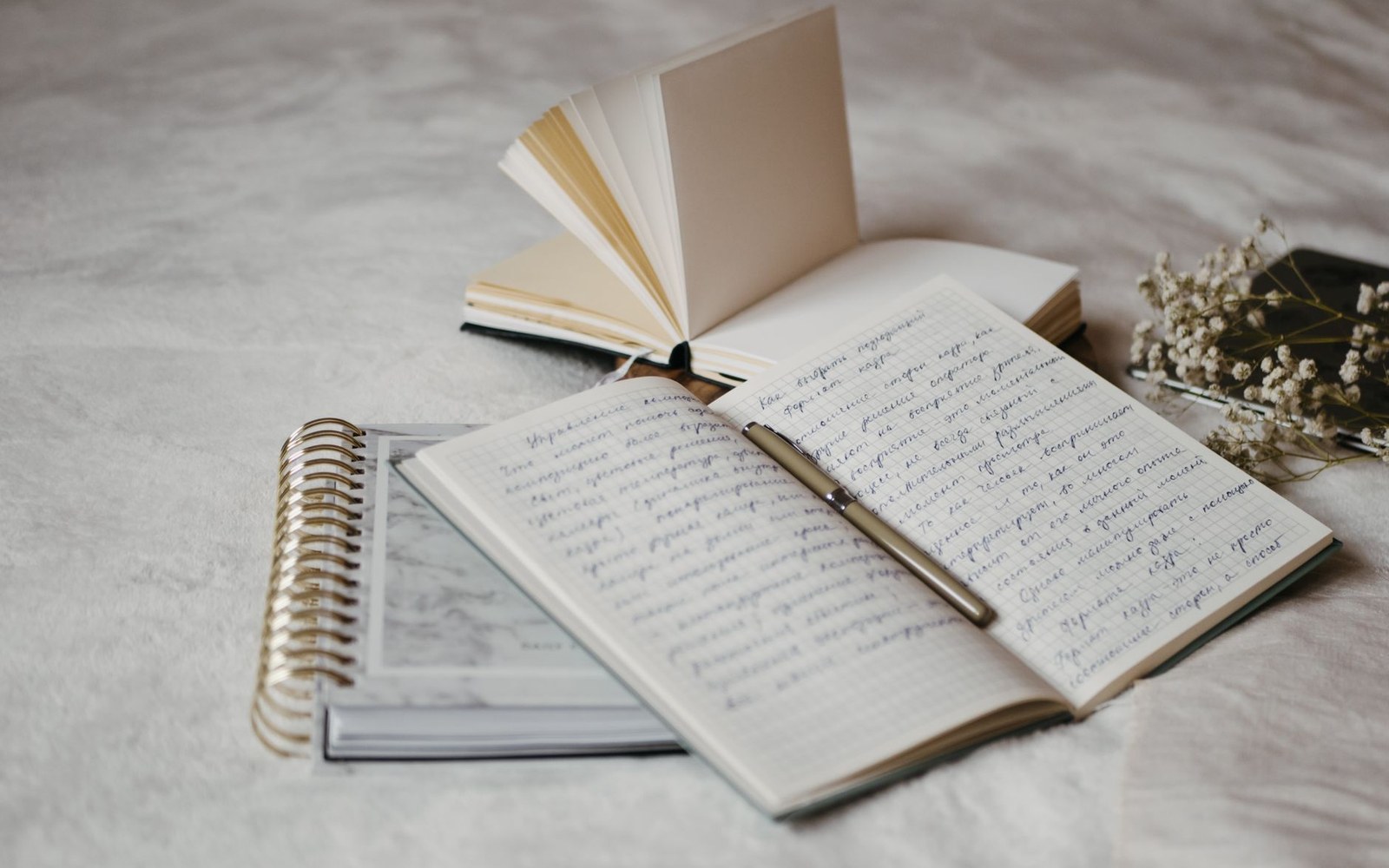 Open handwritten notebook with a pen resting on the page, near a spiral notepad and dried flowers