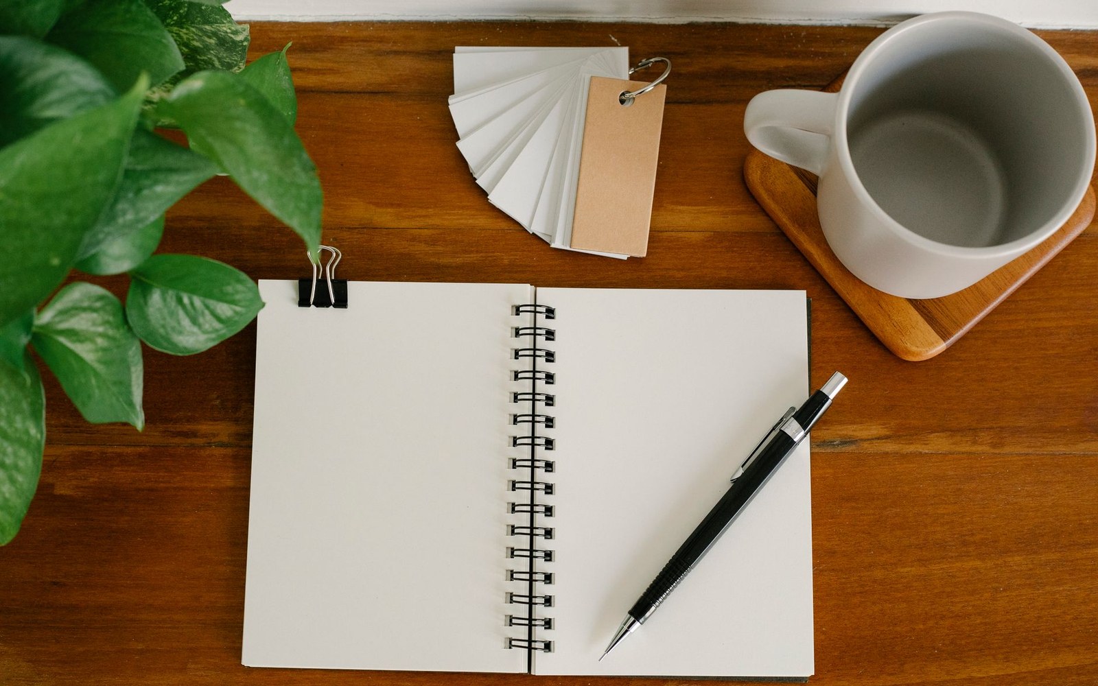 An open spiral notebook, pen, mug, and index cards on a wooden desk
